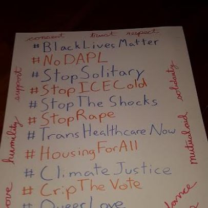 A sign with a bunch of hashtags in the middle:#BlackLivesMatter; #No DAPL; #StopSolitary; #StopICECold; #StopTheShocks; #StopRape; #TransHealthcareNow; #HousingForAll; #ClimateJustice; #CropTheVote; #QueerLove; #resist. Various words in cursive surround the hashtags, including love, humility, support, trust, outrage, mutual aid, solidarity, interdependence, compassion, and courage.