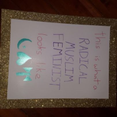 A sign with gold glitter around the border and the text "this is what a RADICAL MUSLIM FEMINIST looks like." A crescent and star, heart pointing up, and labrys are drawn in green at the bottom.