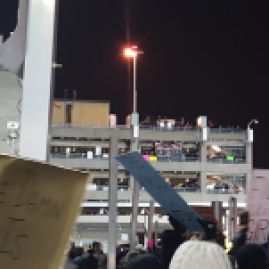 A photo of people protesting. Protesters are both in the foreground at ground level and packing levels of a parking garage. the most prominent banner in the parking garage reads "No Borders"