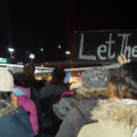 A picture of a bunch of people dressed warmly protesting. The most prominent sign reads "Let Them In"
