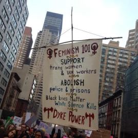 A picture of people marching down a city street with a big sign saying: "Feminism 101: Support trans women and sex workers, Abolish police and prisons, Black lives matter, White silence kills, and Dyke Power